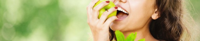 Portrait of young attractive dark-haired smiling woman holding basket full of fruits and eating apple at summer green park.
